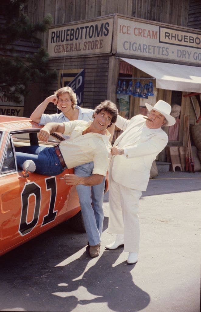 Byron Cherry as Coy Duke leans on a car laughing while Christopher Mayer as Vance Duke is pulled by Sorrell Booke as Boss Hogg outside Rhuebottoms General Store.