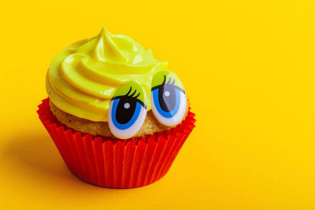 A cupcake in a red paper case topped with bright green swirled frosting and two large cartoon googly eyes, set against a yellow background.