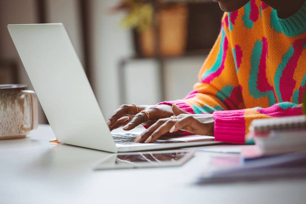 Afro american young woman wearing colorful sweater sitting at the desk at home and using laptop. Close up of hands, unrecognizable person.