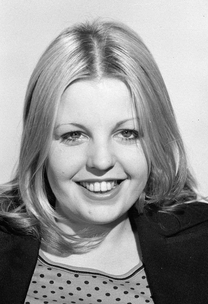 A black and white close-up portrait of a young woman with long, side-parted hair, smiling broadly at the camera while wearing a polka-dot top under a dark jacket.