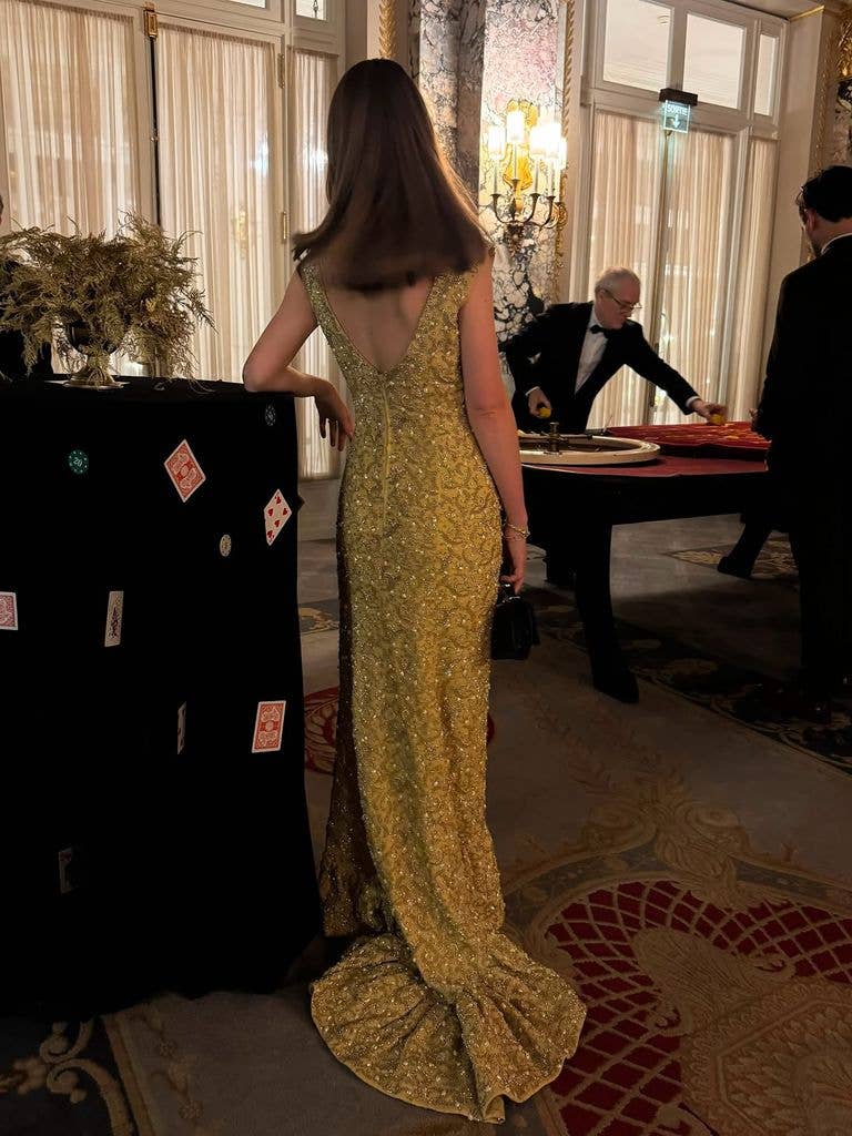 Woman in gold gown stands by table with cards and flowers, two men in suits nearby.