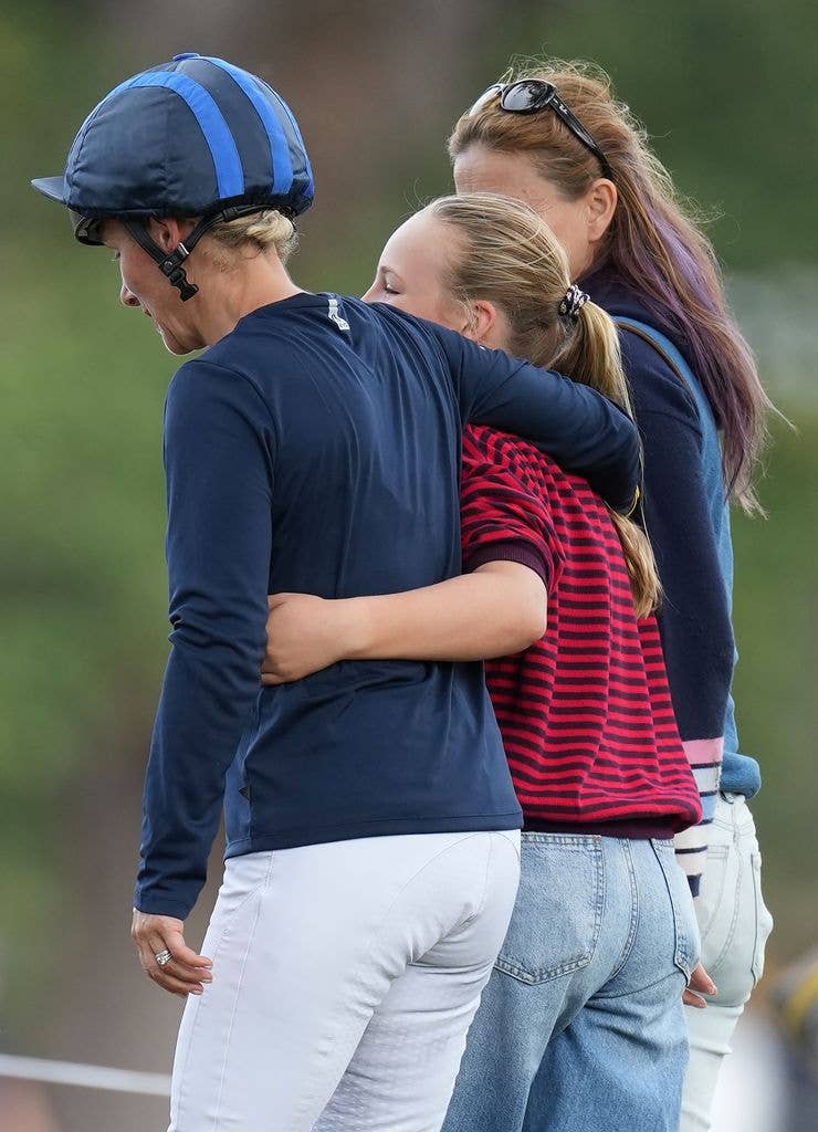 Zara Tindall, wearing a riding helmet and navy top, is embraced from behind by her daughter Mia Tindall as another woman stands nearby, with green parkland in the background.