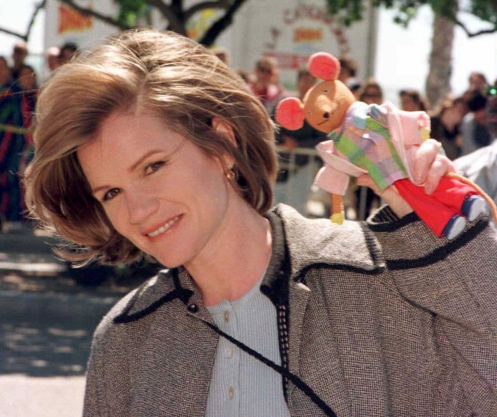 Actress and Oscar nominee Mare Winningham sholds her head at an angle down towards the left of the image, her hair in a short, side parted bob. She wears a grey buttoned cardigan and a brown grey jacket and shows her daughter's toy mouse in a checked shirt, pink jacket and red trousers. 