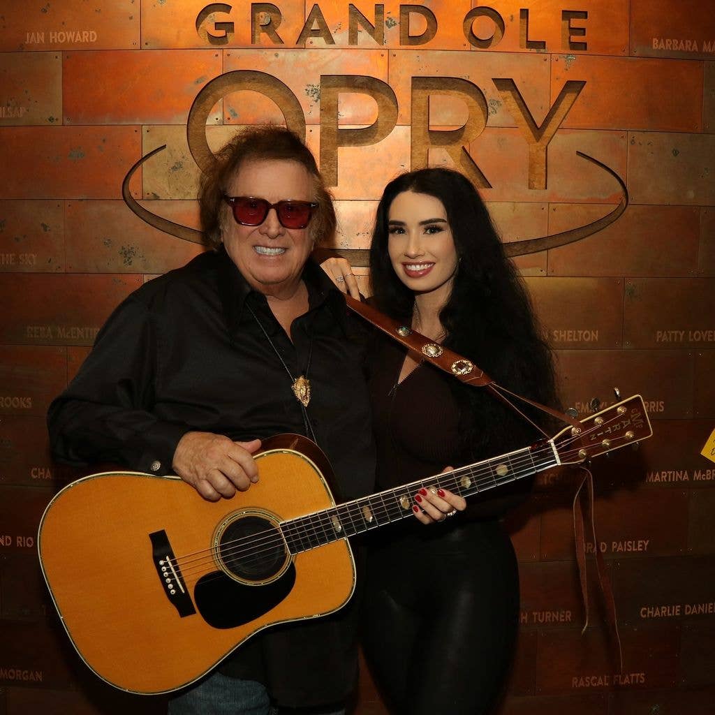 Don McLean, wearing sunglasses and holding an acoustic guitar, smiles alongside a woman with long dark hair in front of the Grand Ole Opry sign.