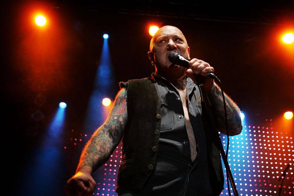 Angry Anderson of Rose Tattoo performs on stage with microphone, tattoos visible.