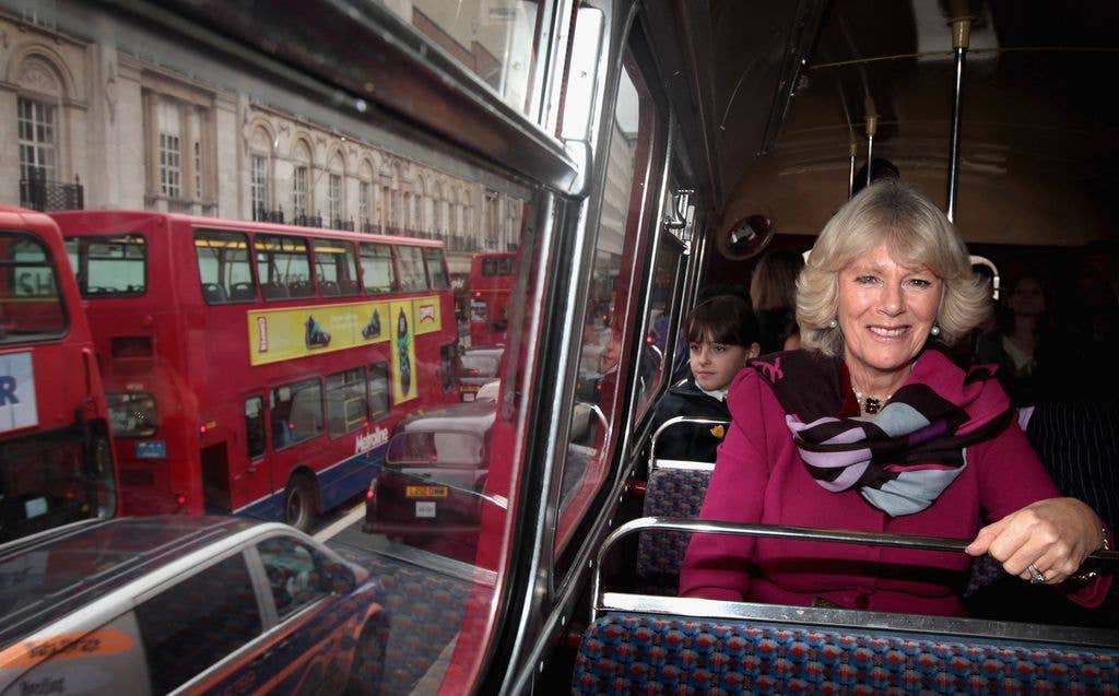 Camilla, Duchess of Cornwall, smiling on a bus with red double-deckers outside.