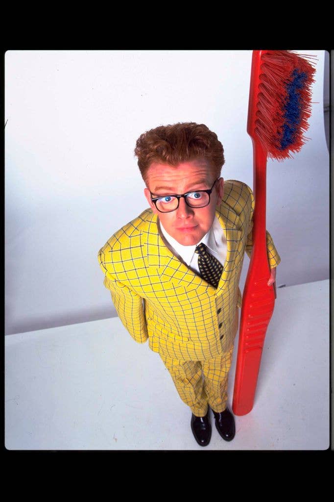 Chris Evans, wearing a yellow checked suit and glasses, stands beside a giant novelty toothbrush, looking up at the camera.