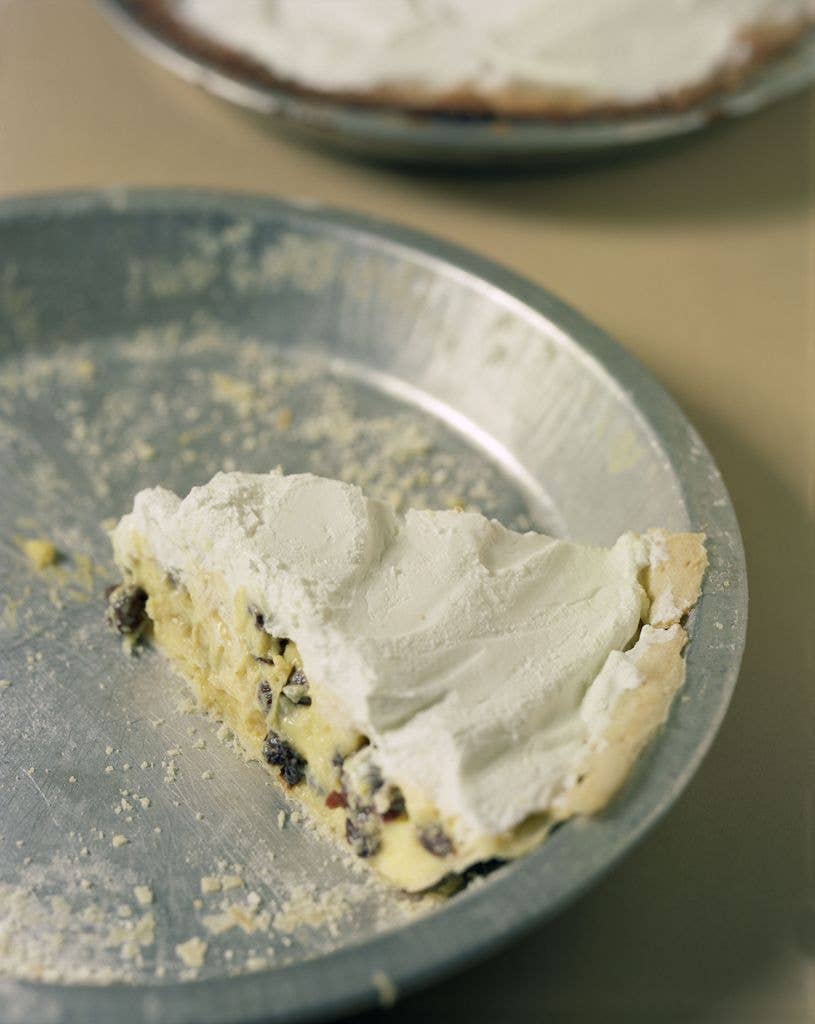 A single slice of sour cream and raisin pie topped with whipped cream sits in a metal pie tin, with a second whole pie visible in the background.