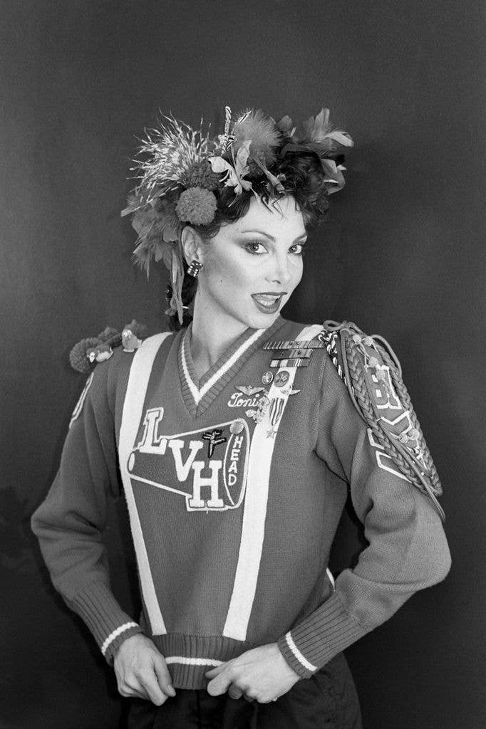 Toni Basil wearing a cheerleader sweater and a feathered headdress, smiling.