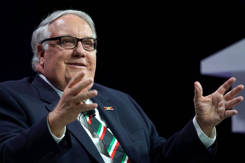 Howard G. Buffett, wearing glasses and a striped tie, gestures with both hands while speaking on stage against a dark background.