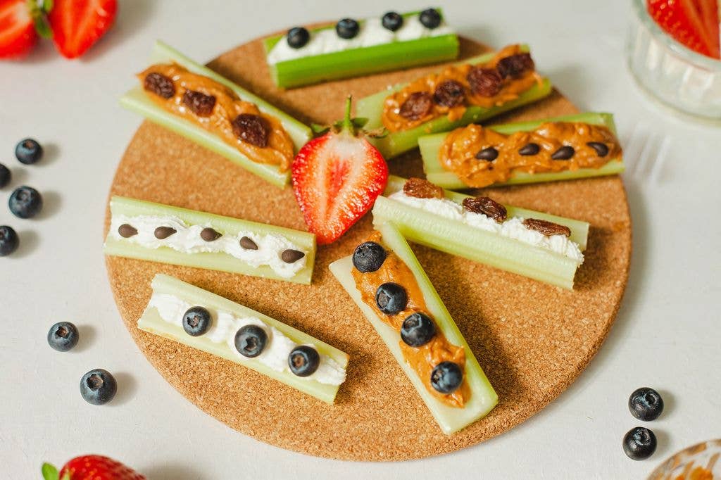 Celery sticks arranged in a circle on a wooden board, filled with peanut butter or cream cheese and topped with raisins, blueberries or chocolate chips, with strawberries and blueberries scattered nearby.