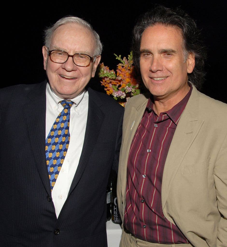 Warren Buffett, wearing a dark suit and patterned tie, smiles next to his son Peter Buffett, dressed in a striped shirt and beige jacket, at an event.
