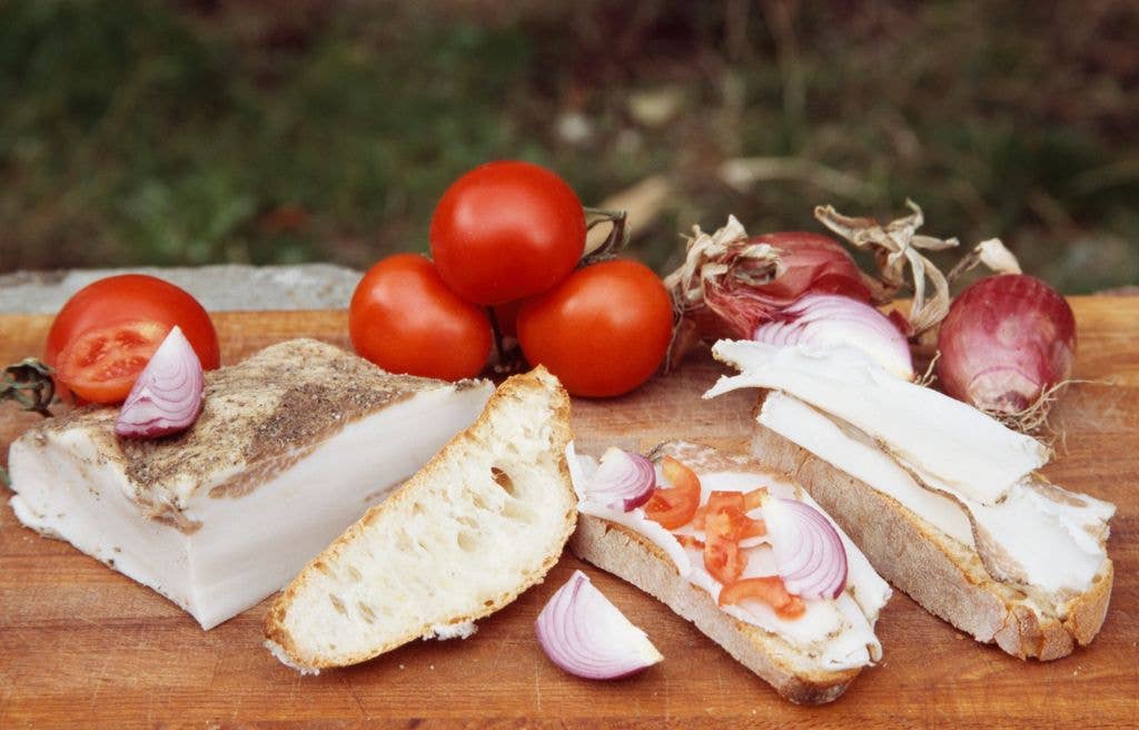 A block of cured white lard sits on a wooden board alongside slices of bread topped with lard, tomato and red onion, with whole tomatoes and onions in the background.