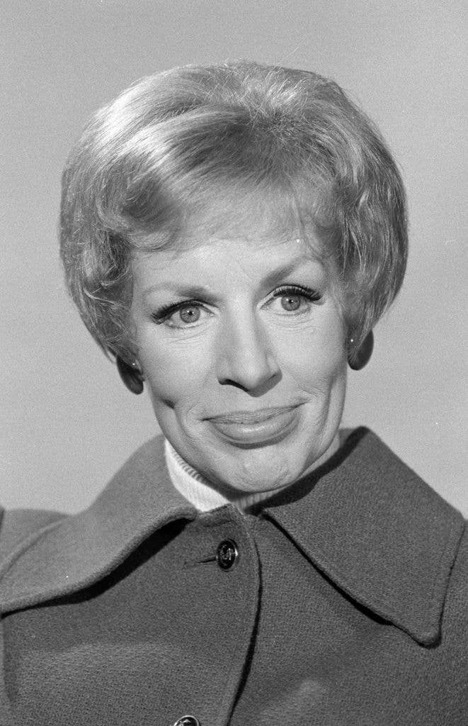 A black-and-white close-up portrait of a woman with short, styled hair, wearing false eyelashes and a coat with a large collar, smiling gently at the camera.