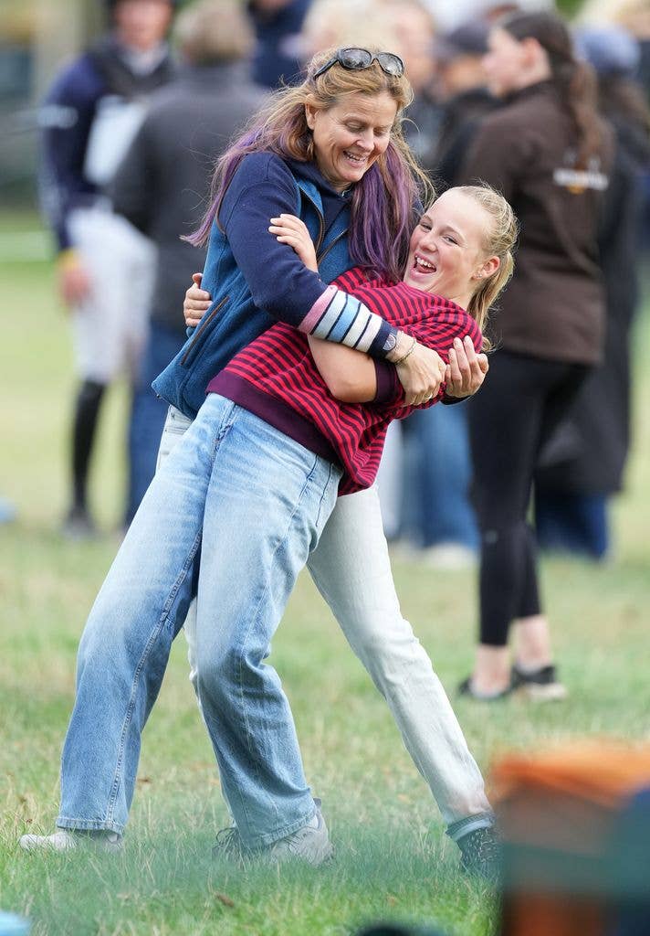 A woman embraces Mia Tindall, both laughing, at an outdoor equestrian event with onlookers in the background.