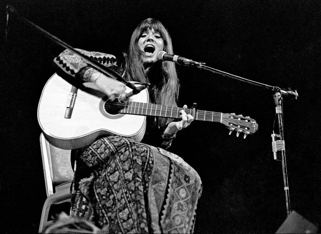 Black and white photograph of Melanie singing into a microphone while playing acoustic guitar, her long hair falling forward as she performs on stage.
