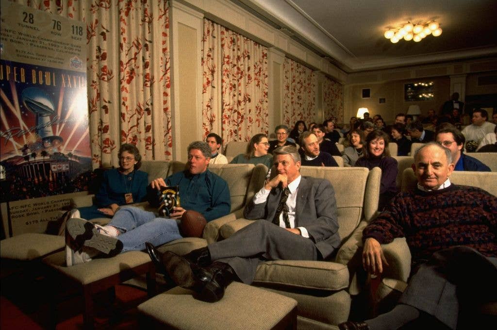 President Bill Clinton (C) watching Super Bowl in family theater with governor guests and White House staffers, munching popcorn, with sneakered feet on ottoman and a football at his side in 1994