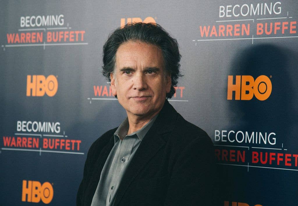 Peter Buffett stands on a step-and-repeat backdrop printed with the HBO and Becoming Warren Buffett logos, wearing a dark jacket and grey shirt.