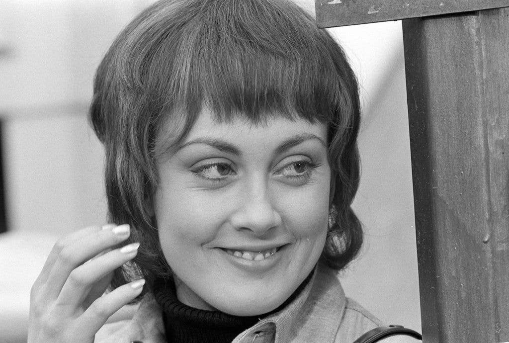 A black and white close-up photograph of a young woman with a fringed bob hairstyle, smiling and touching her hair near her face.