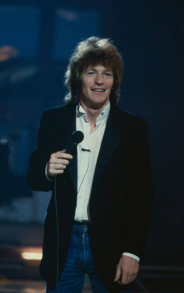 Jim Davidson stands on a dimly lit stage holding a microphone, smiling, wearing an open white shirt, dark blazer and jeans.