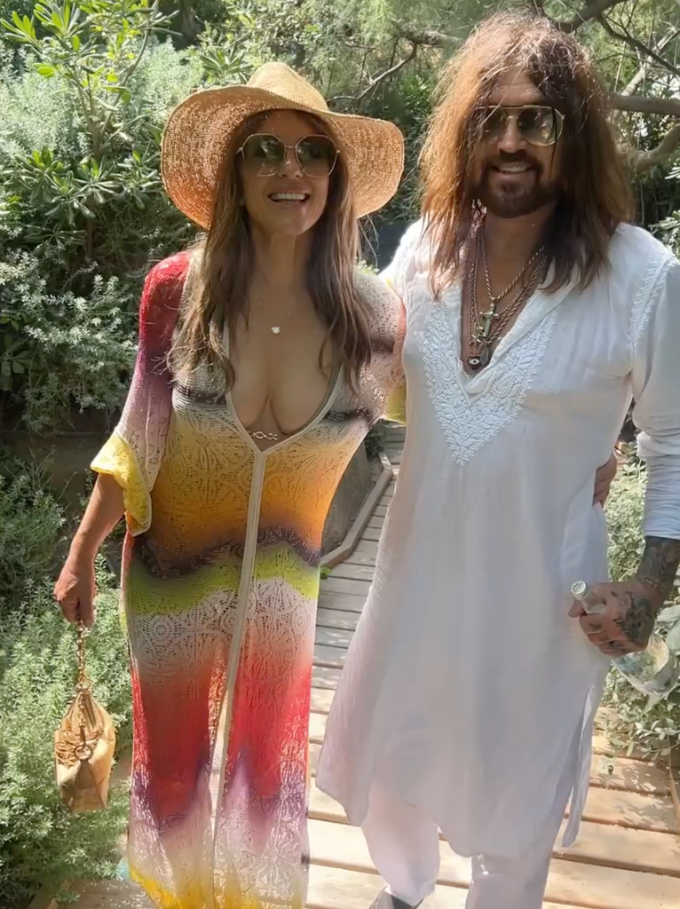 Elizabeth Hurley and a man with long hair and sunglasses stand together on a wooden path, smiling.
