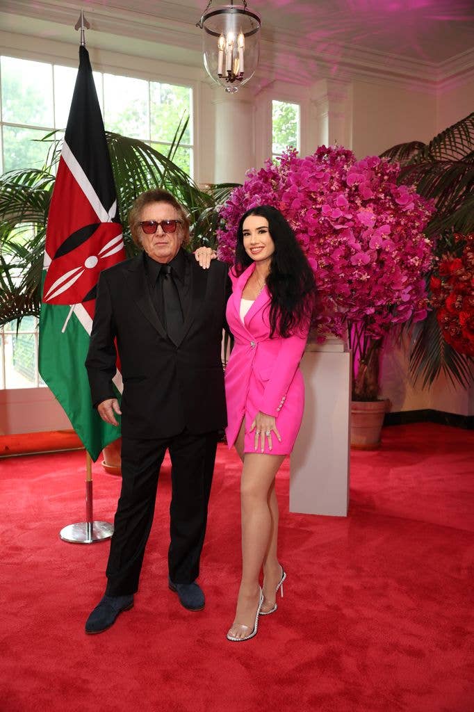 Don McLean in a black suit and sunglasses poses with Paris Dylan in a hot pink suit on a red carpet near a Kenyan flag and pink orchid display at the White House.