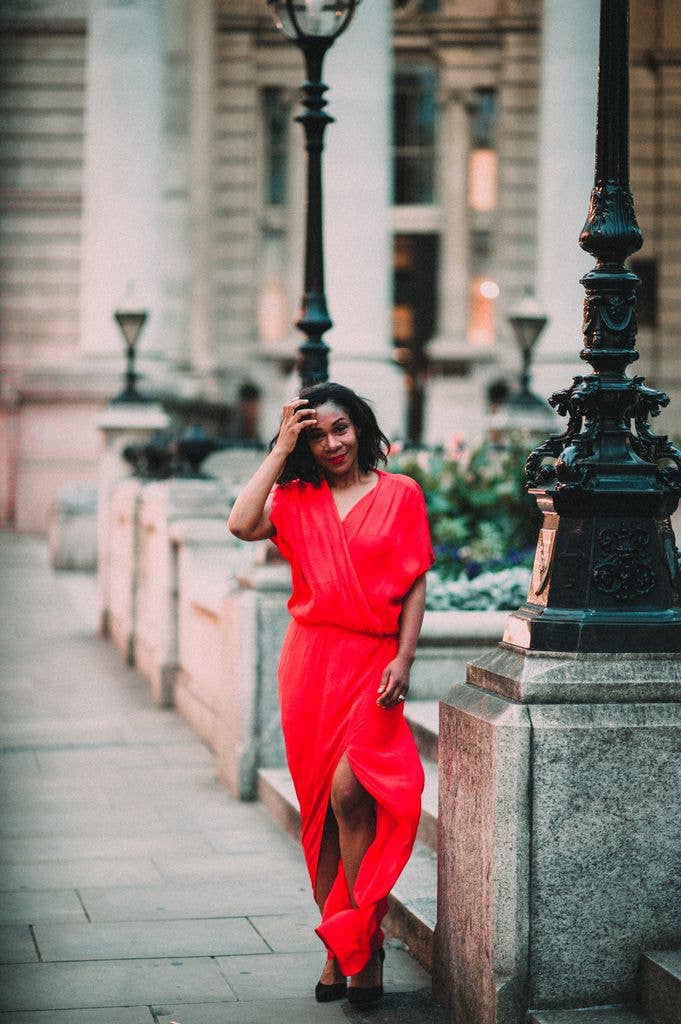 Karen Bryson in a red wrap dress stands on a city sidewalk, hand on her head.