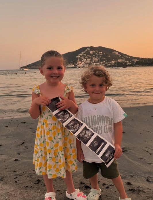 Two children on a beach holding an ultrasound scan. The girl wears a lemon print dress, and the boy wears a white t-shirt with 'Millionaire's Club' text.